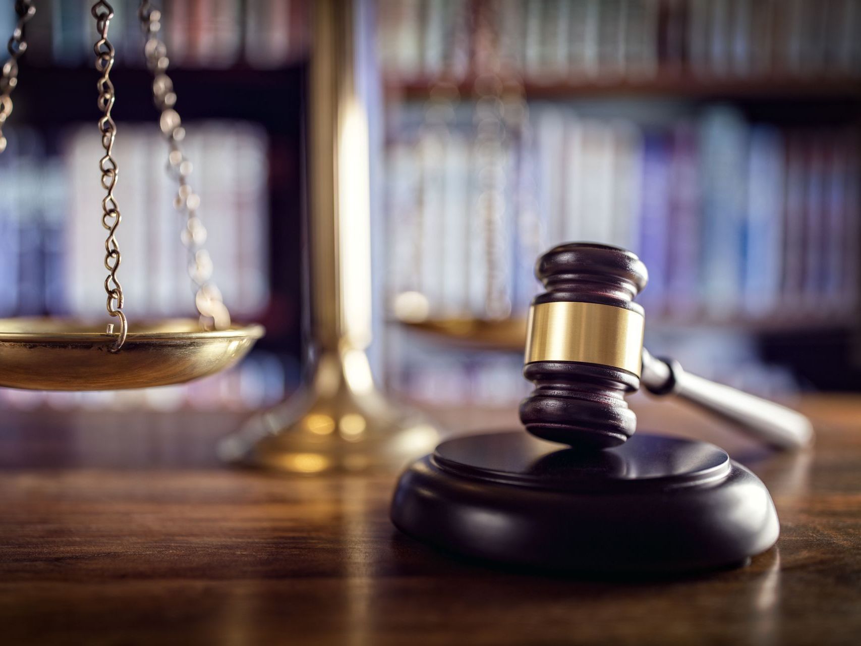 A wooden judge's gavel rests on a desk in front of a blurred set of scales of justice and a law library bookshelf.