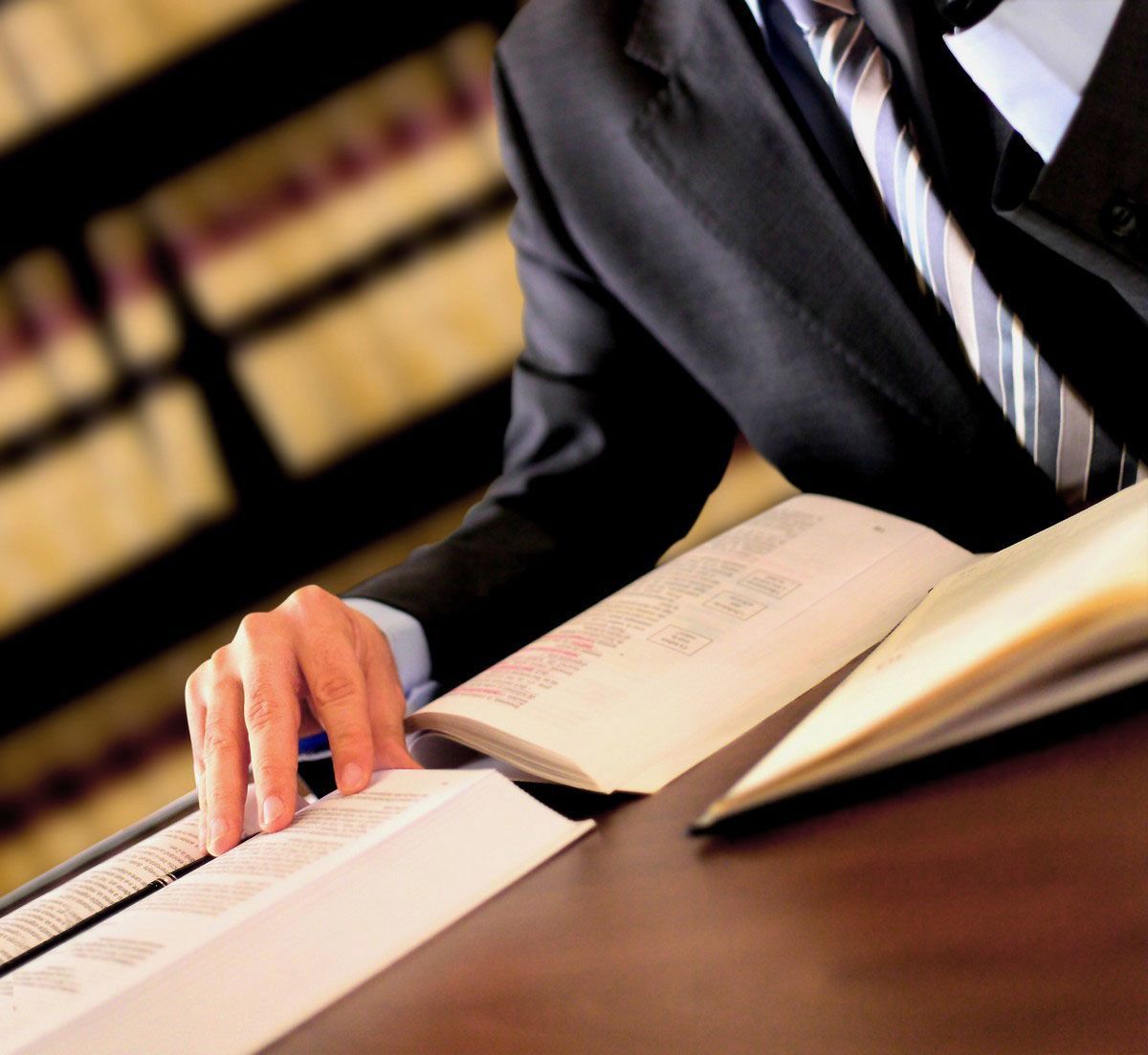 A person in a suit and tie reads a legal text in a law library with shelves of books in the background.