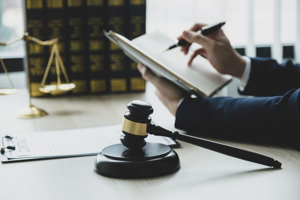 A wooden gavel sits on a desk in front of a legal professional writing in a book, with law books and a scale in the back.