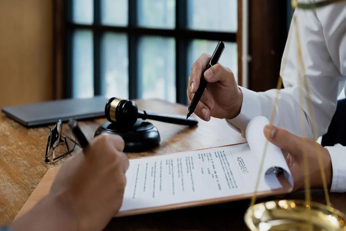 Two people signing legal documents on a desk with a judge's gavel and a scale of justice nearby.