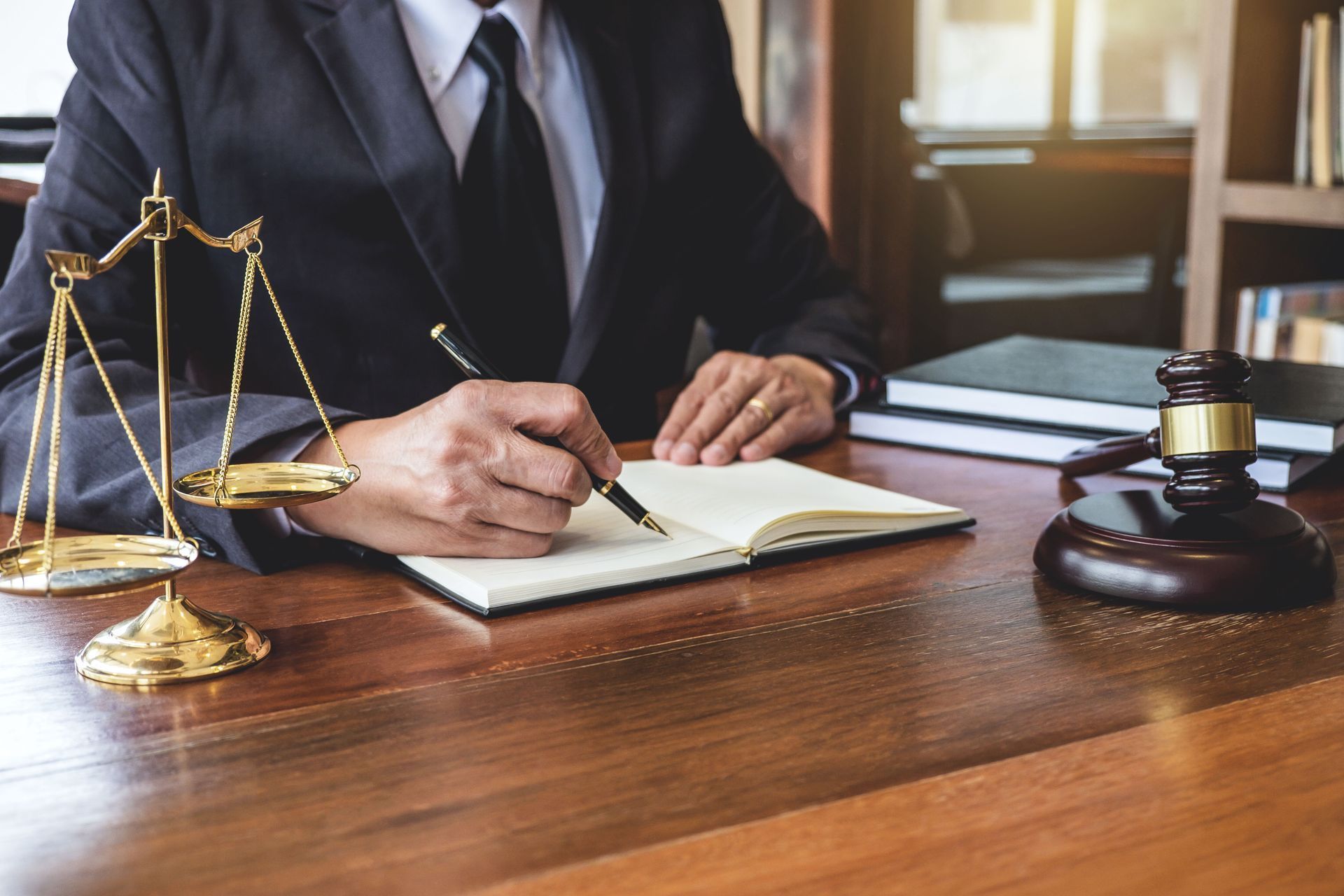 A person in a suit writes in a notebook at a desk featuring a pair of scales and a wooden gavel.
