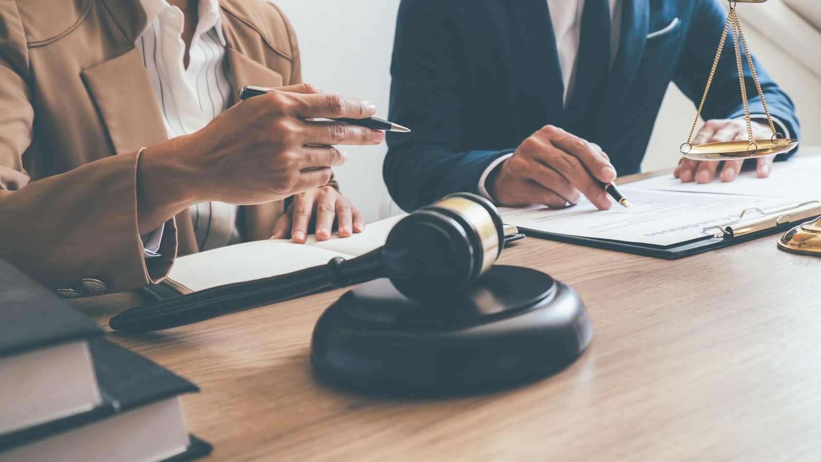 A wooden gavel on a desk with two professionals reviewing legal documents and a scale of justice in the background.