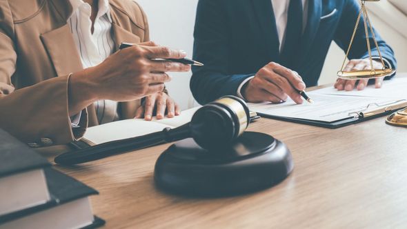 A wooden gavel on a desk with two professionals reviewing legal documents and a scale of justice in the background.