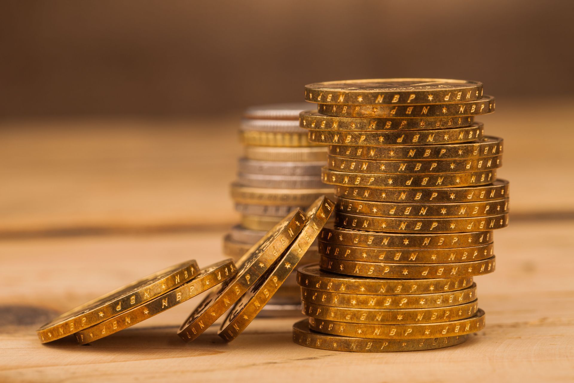 A stack of gold coins sits on a wooden surface, with several loose coins leaning against its side.