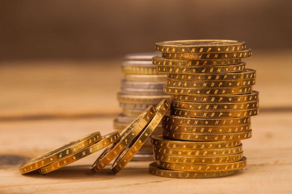 A tall stack of gold coins sits on a wooden surface next to a smaller, slanted pile of gold coins.