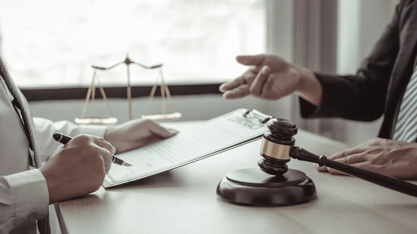 Two people seated at a desk with a gavel and scales of justice, one signing a document and the other gesturing.