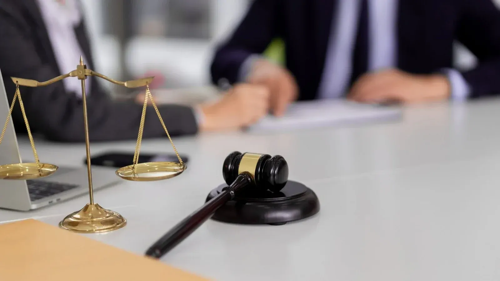 A wooden gavel and brass scales of justice sit on a desk in front of blurred figures in business attire.