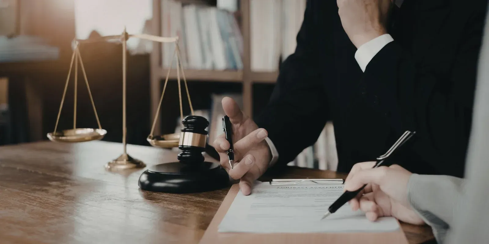 A lawyer’s desk featuring a brass scale of justice, a wooden gavel, and two people reviewing legal documents.