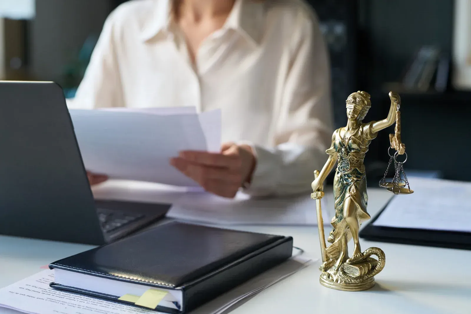 A person in a white shirt sits at a desk with a laptop and papers, next to a gold statue of Lady Justice.