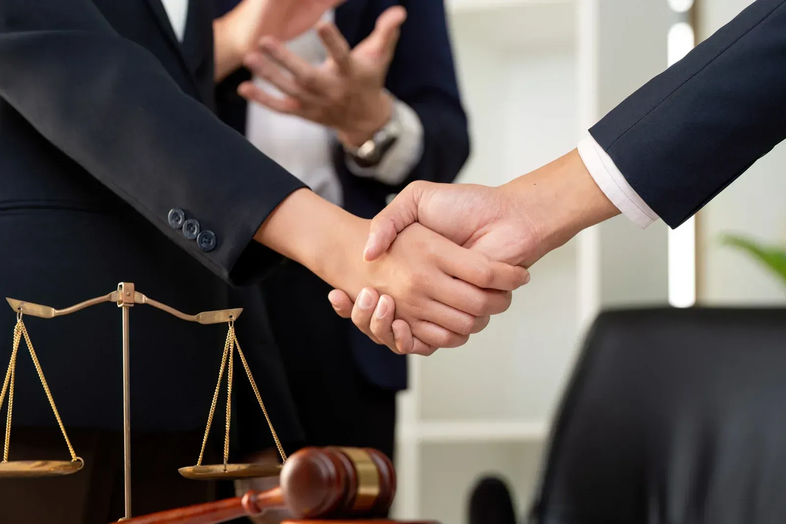 Two people in business suits shake hands in front of a law scale and gavel, signifying a legal agreement.