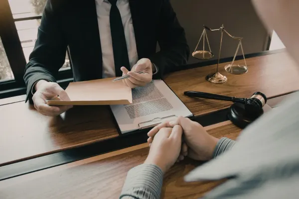 A lawyer in a suit presents a document to a client across a wooden desk with a gavel and balance scale nearby.
