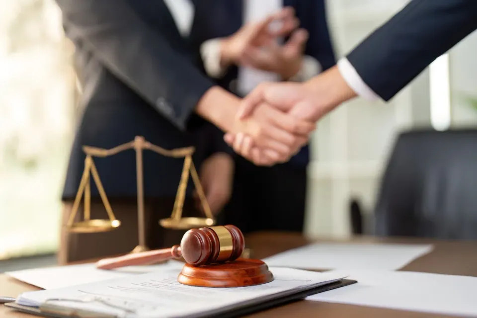 Two people in business suits shake hands over a legal document, gavel, and scales of justice on a desk.