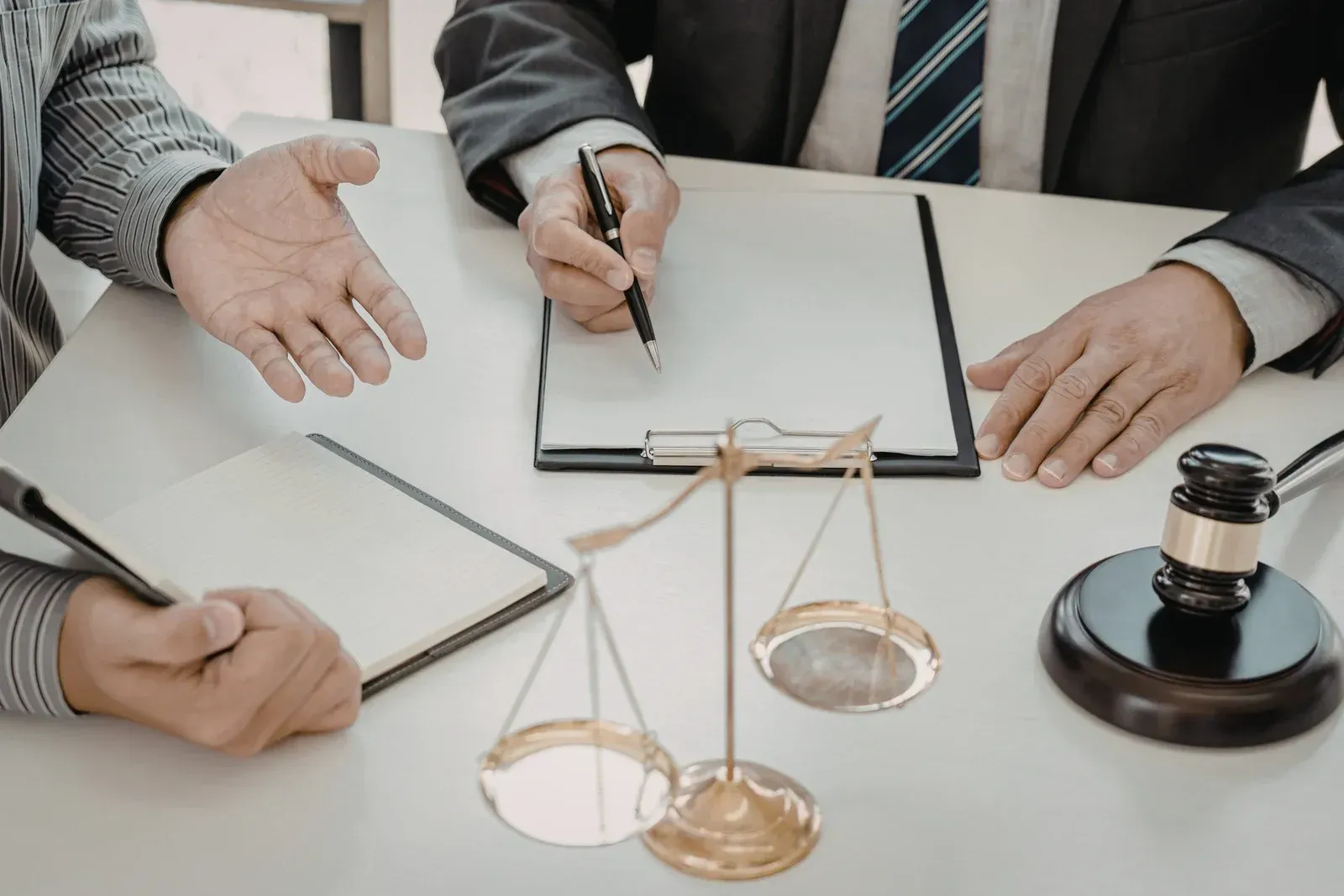 Two people sit at a desk with legal documents, a pen, and a judge's gavel beside a gold scale of justice.