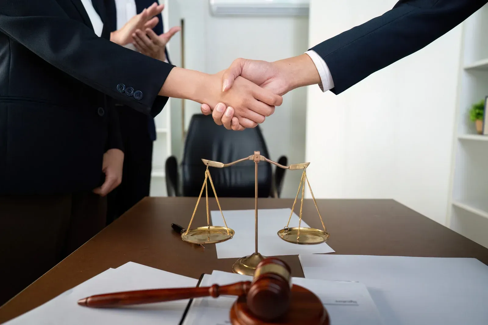 Two professionals in suits shake hands over a wooden table with a brass scale of justice and a wooden judge’s gavel.