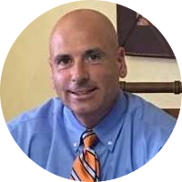 Bald man in blue shirt, orange tie, smiling, indoor setting.