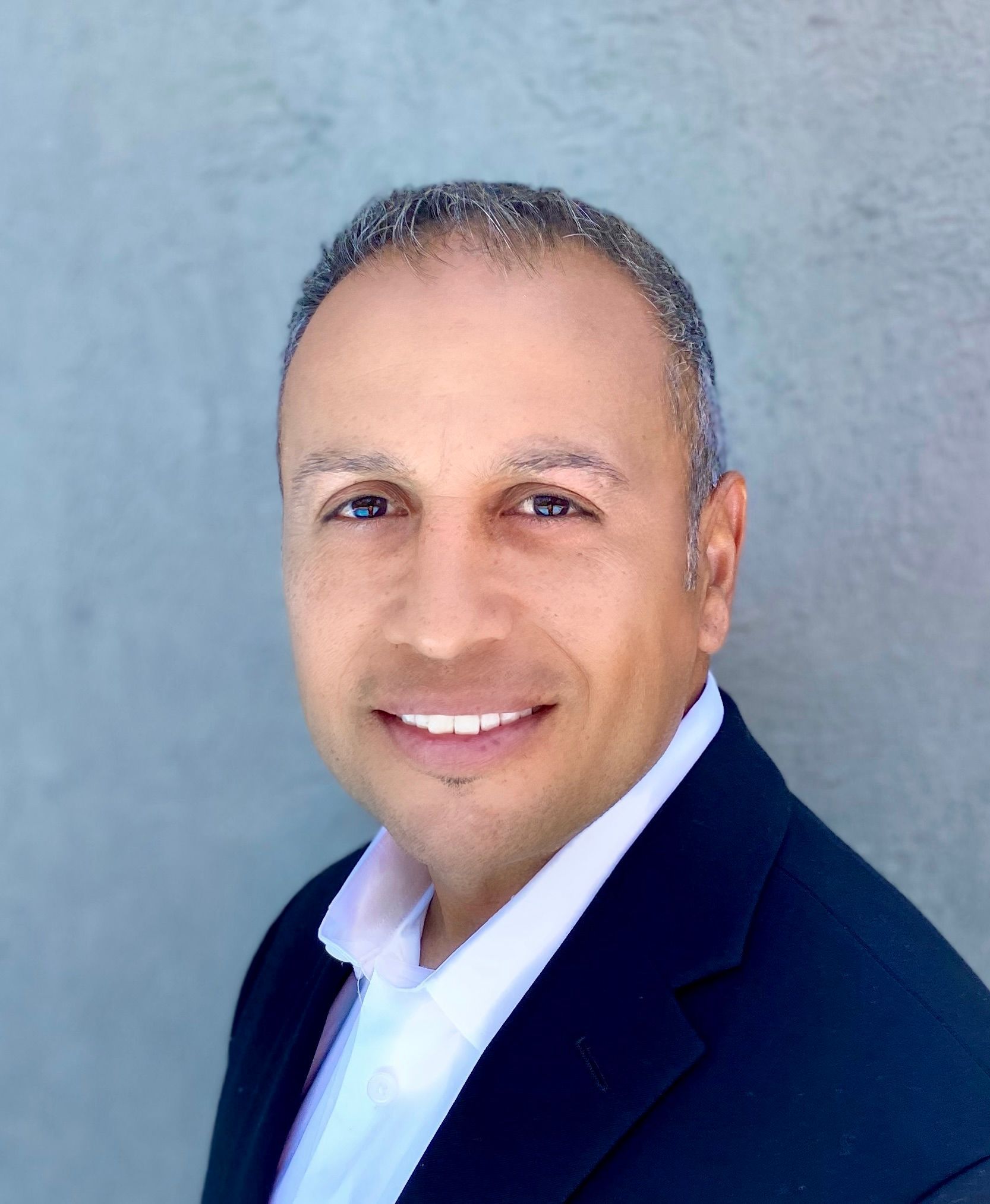Headshot of a smiling person wearing a white collared shirt and a dark navy blazer against a grey background.