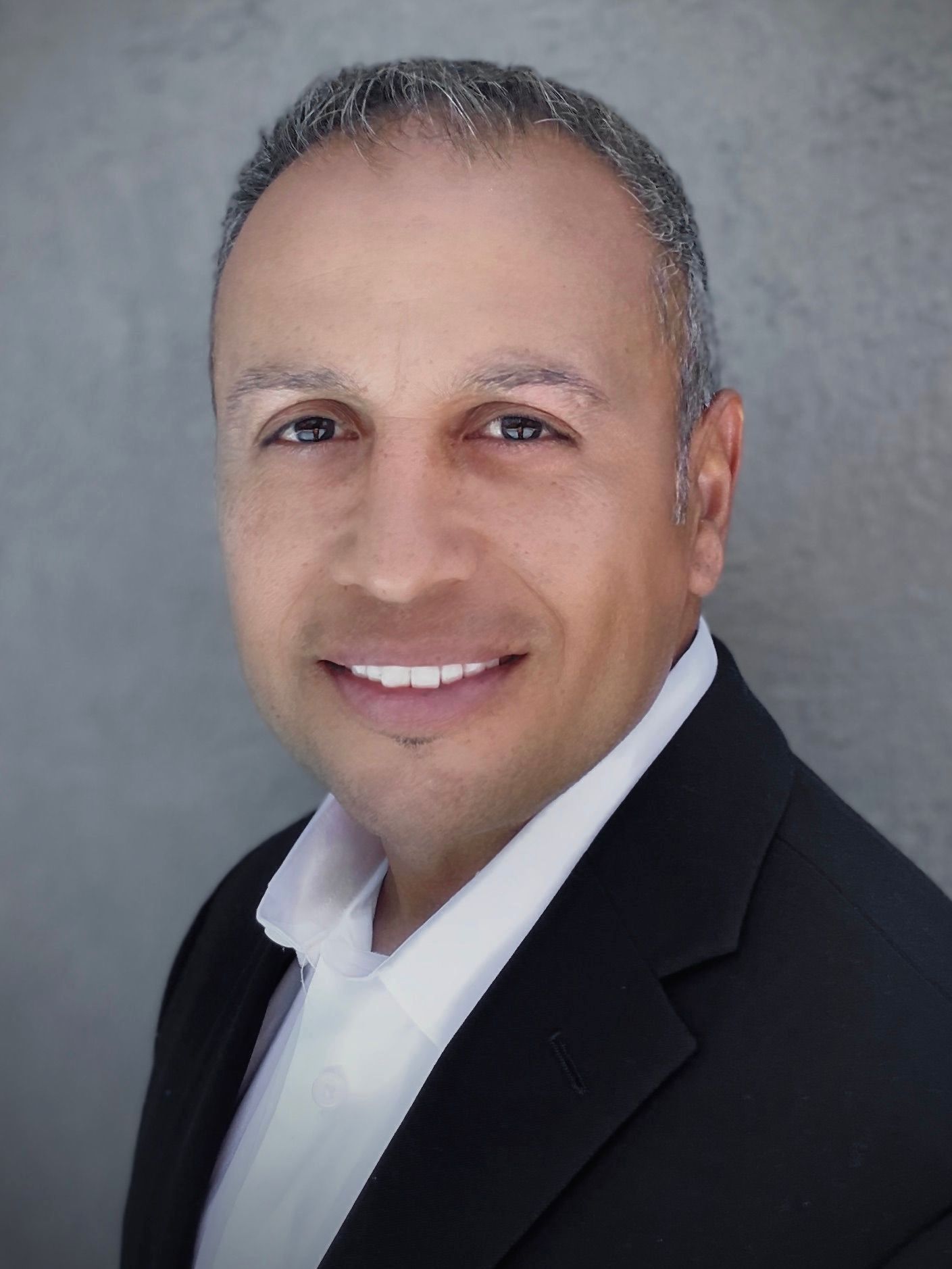 Man in black blazer and white shirt smiles at the camera, neutral grey background.
