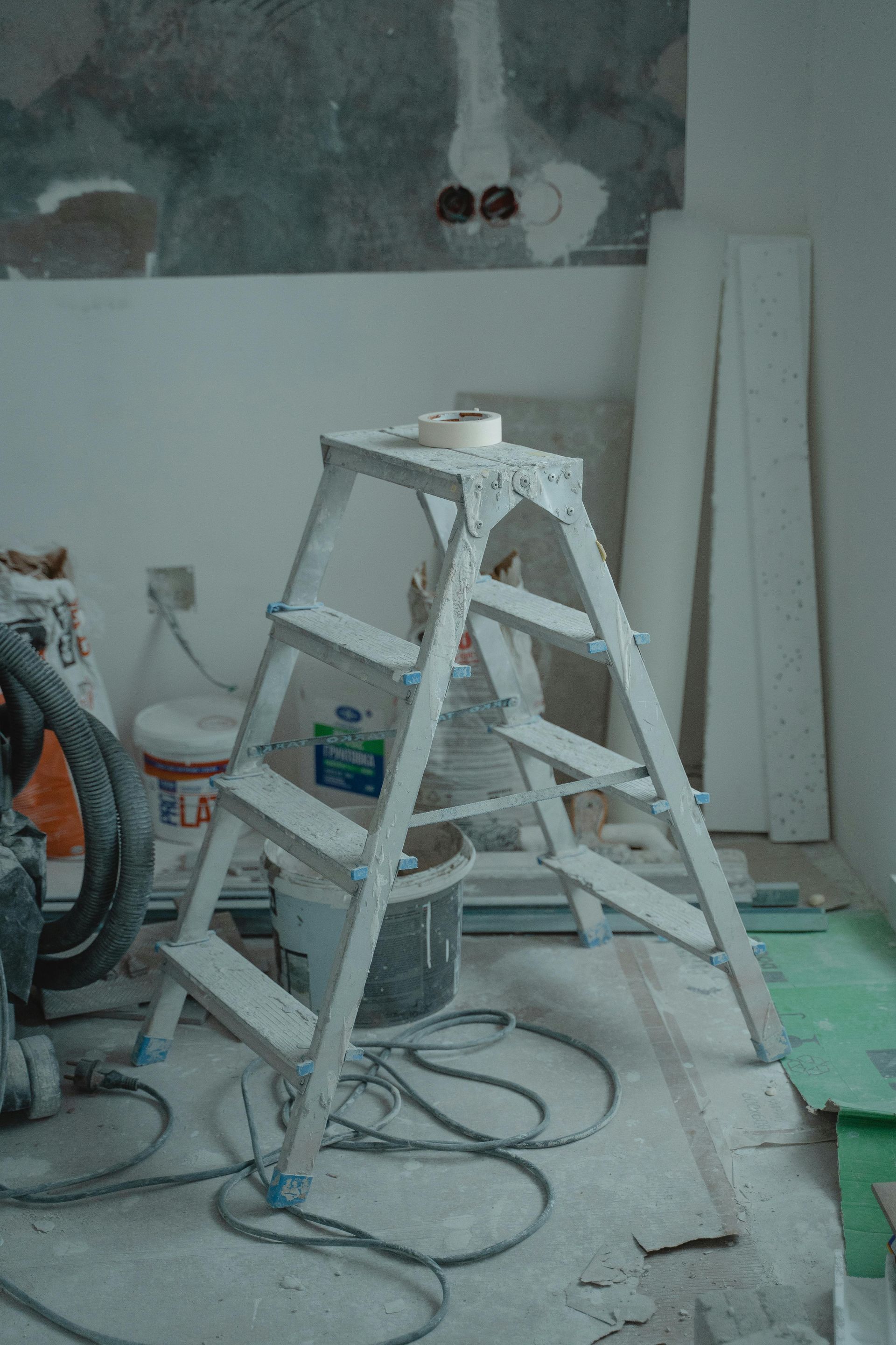 A silver step ladder in a room under construction. There's a bucket, cords, and drywall in the background.