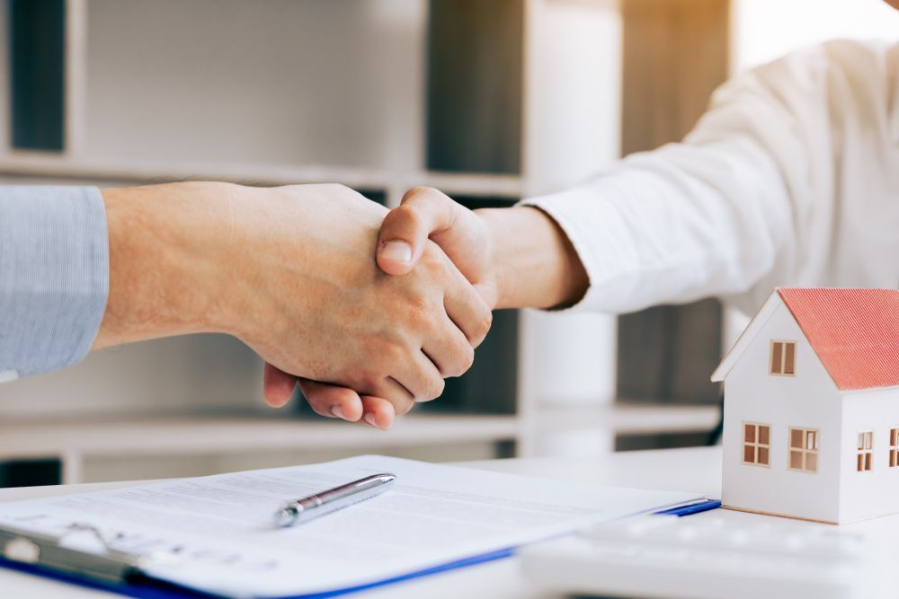 Two People Shaking Hands Over a House Model and Documents — Kellie O'Malley Conveyancing In Lake Munmorah, NSW