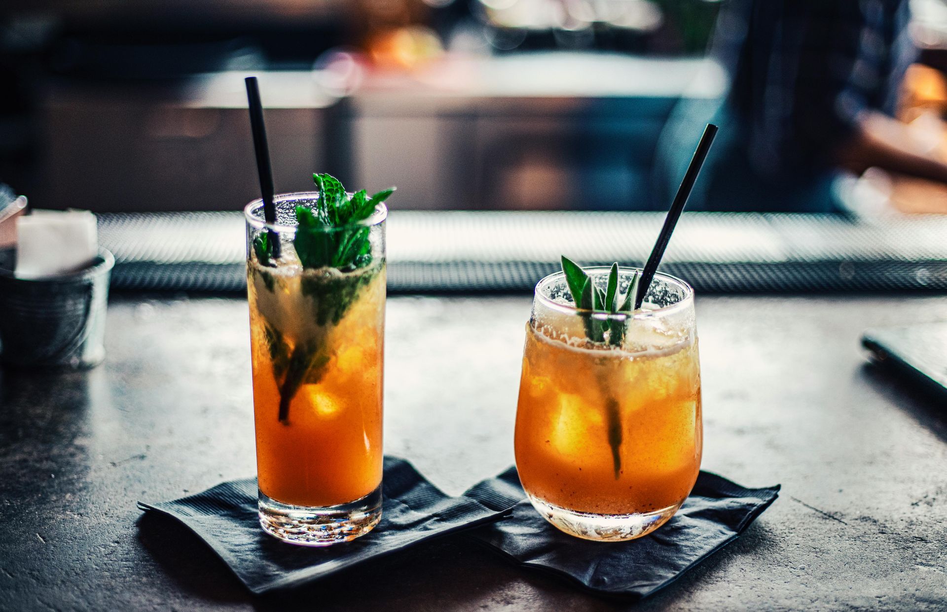 Two cocktails with mint sprigs and straws on a bar, lit with warm light.