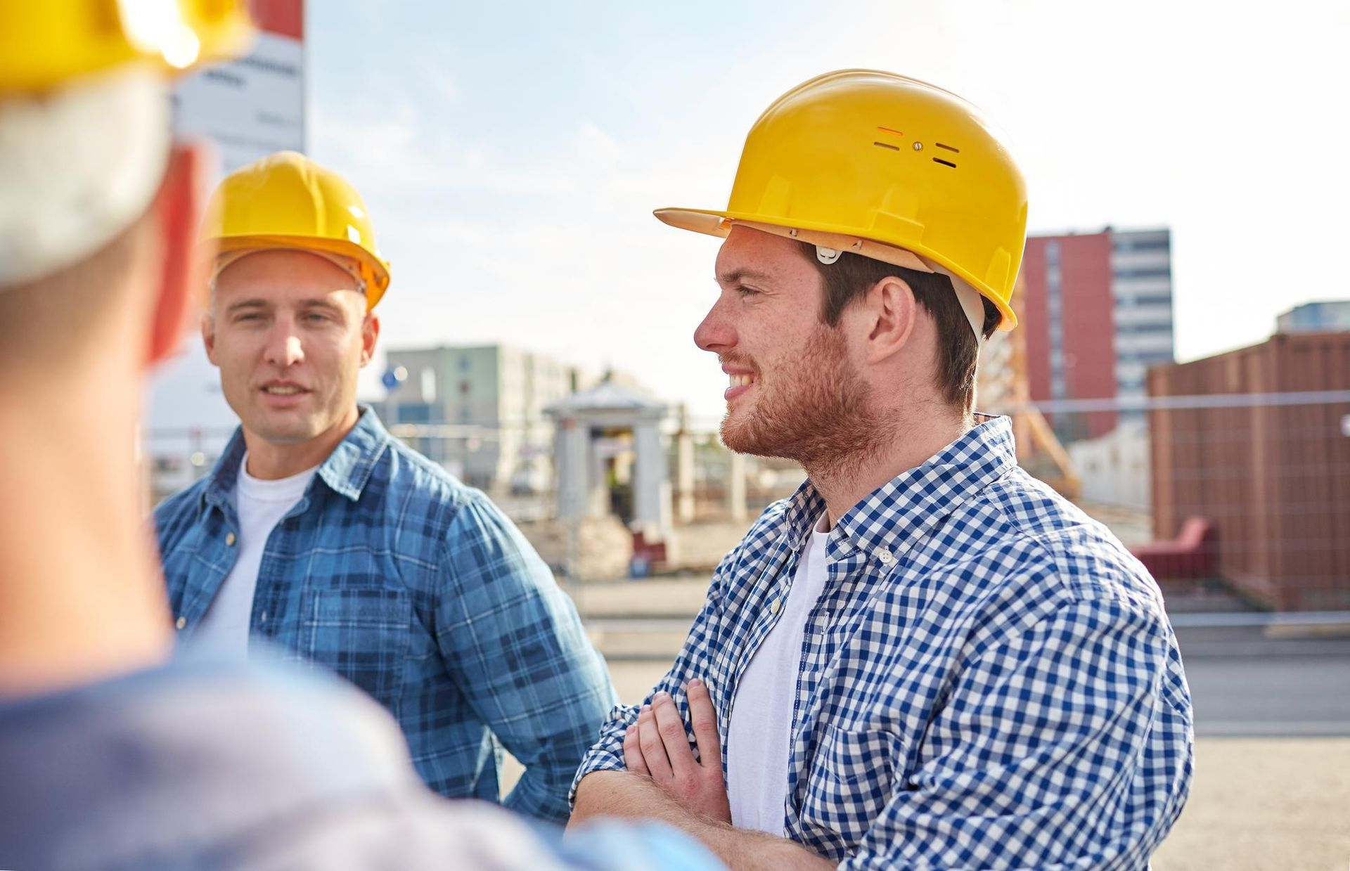 Construction workers in yellow hard hats and blue plaid shirts talking on a sunny construction site.