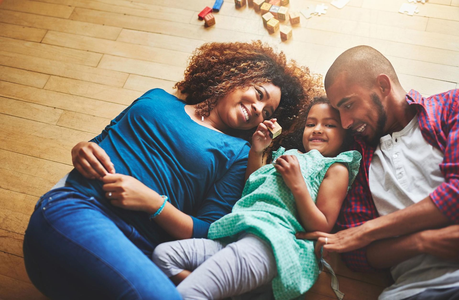 Family of three lying on a wooden floor, smiling and cuddling; toys in background.