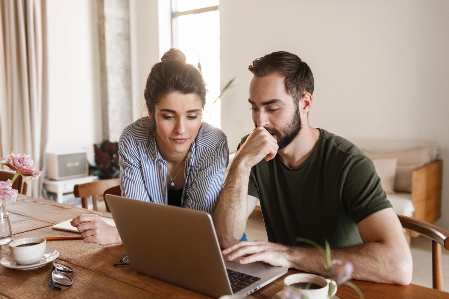 Woman and man looking at a laptop at a table, discussing. Interior setting with coffee, flowers, and window.