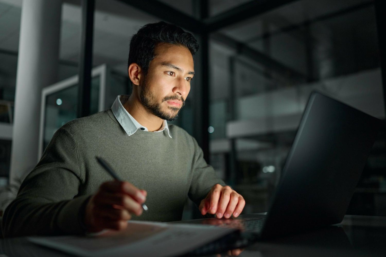Man working on laptop at night, writing on paper in a dimly lit office.