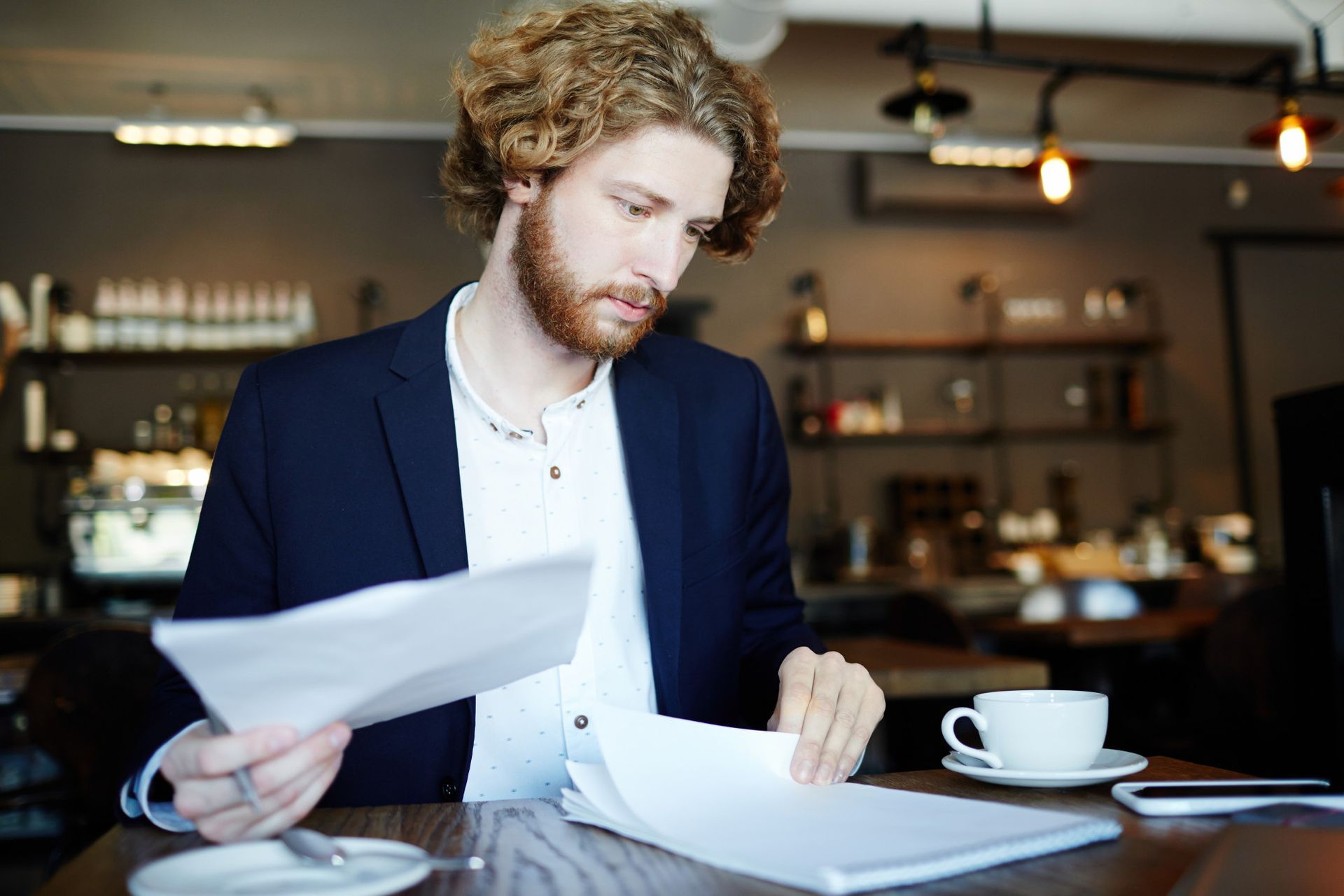 Man in a blazer reviews papers at a table in a cafe, holding documents, with a coffee cup nearby.