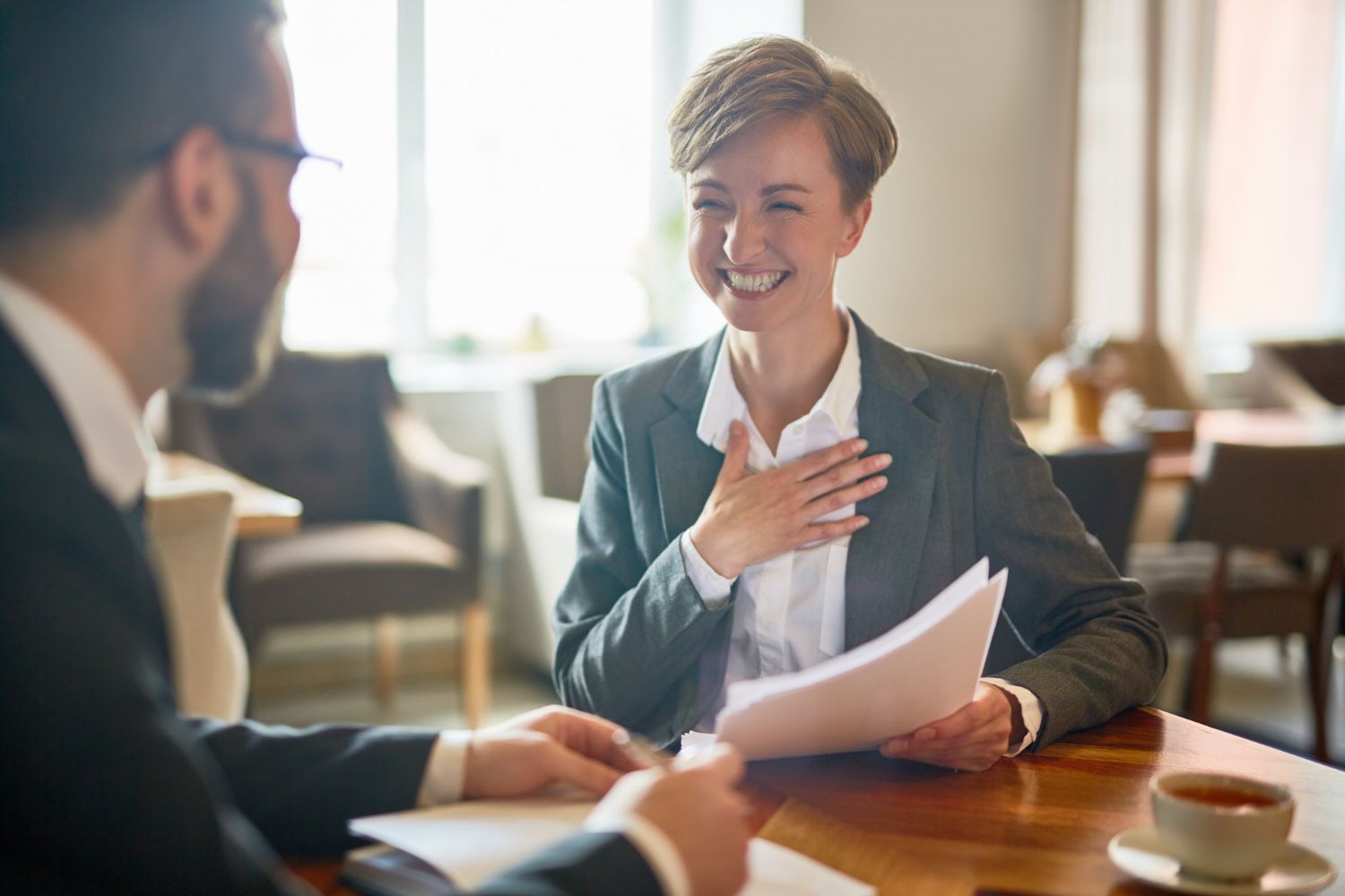Woman in blazer smiles, touches chest, holds papers, seated across from person, table in a cafe.