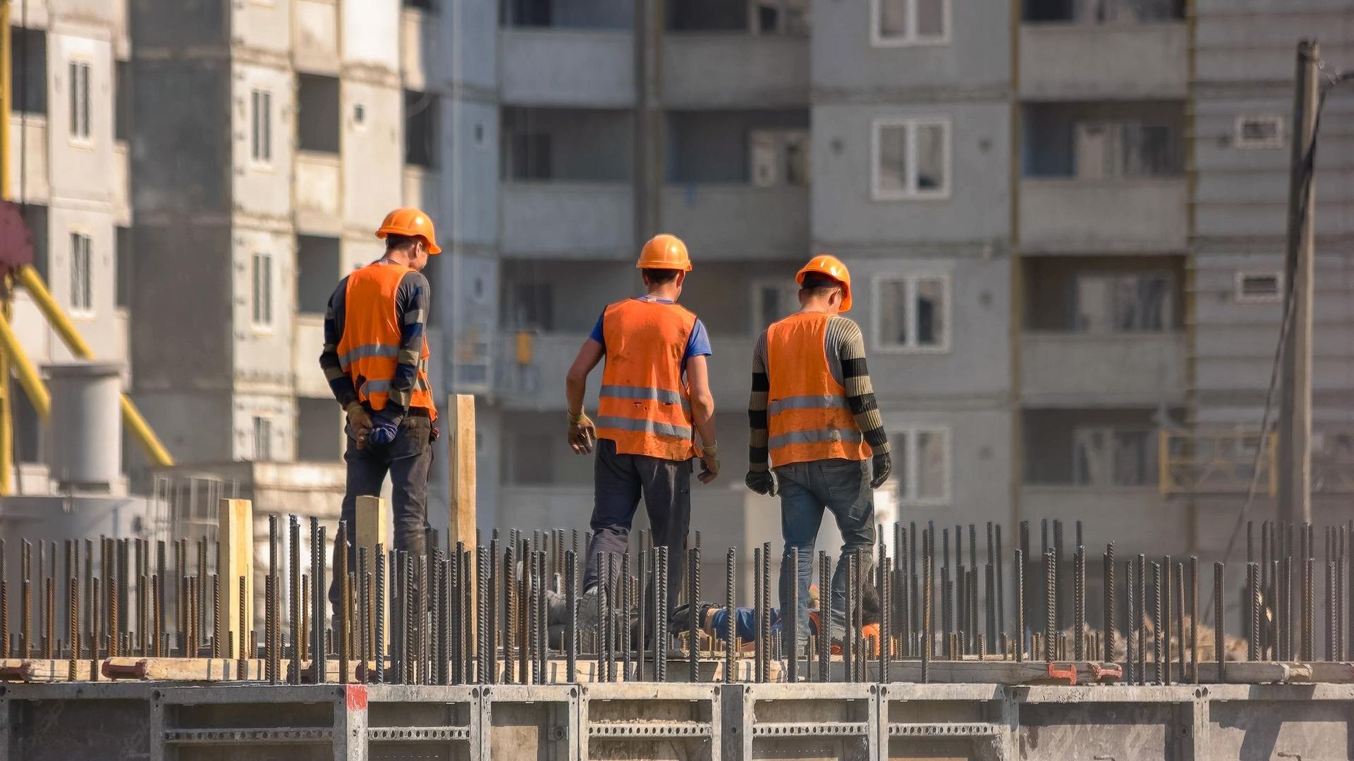 Construction workers on a concrete structure, wearing orange vests and hard hats, with a building in the background.