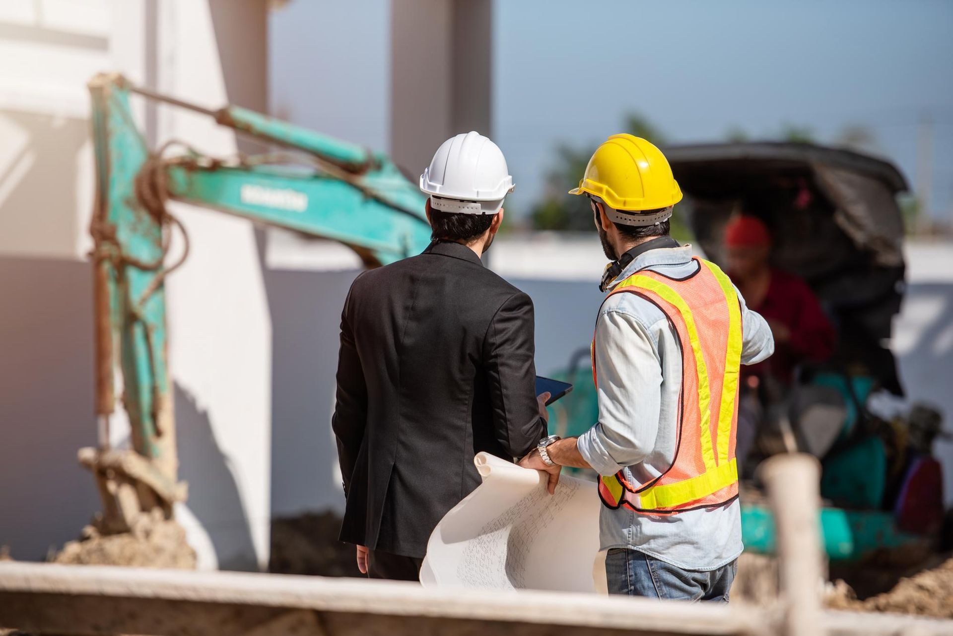 Two men in hard hats reviewing blueprints at a construction site near a small excavator.