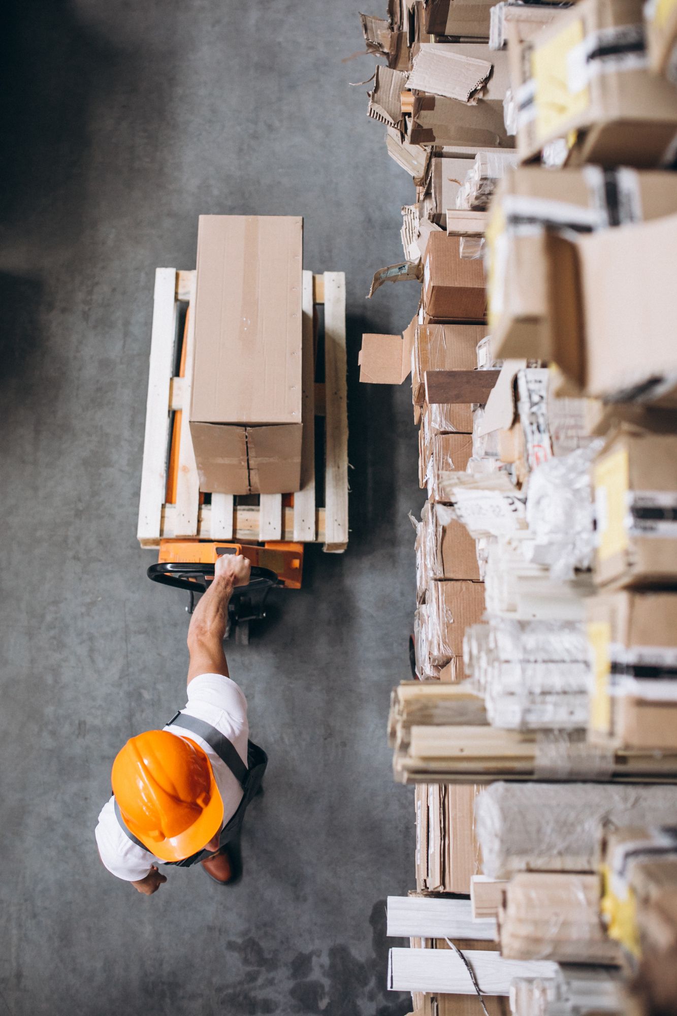 Warehouse worker in orange helmet operating pallet jack, moving boxes near shelves.