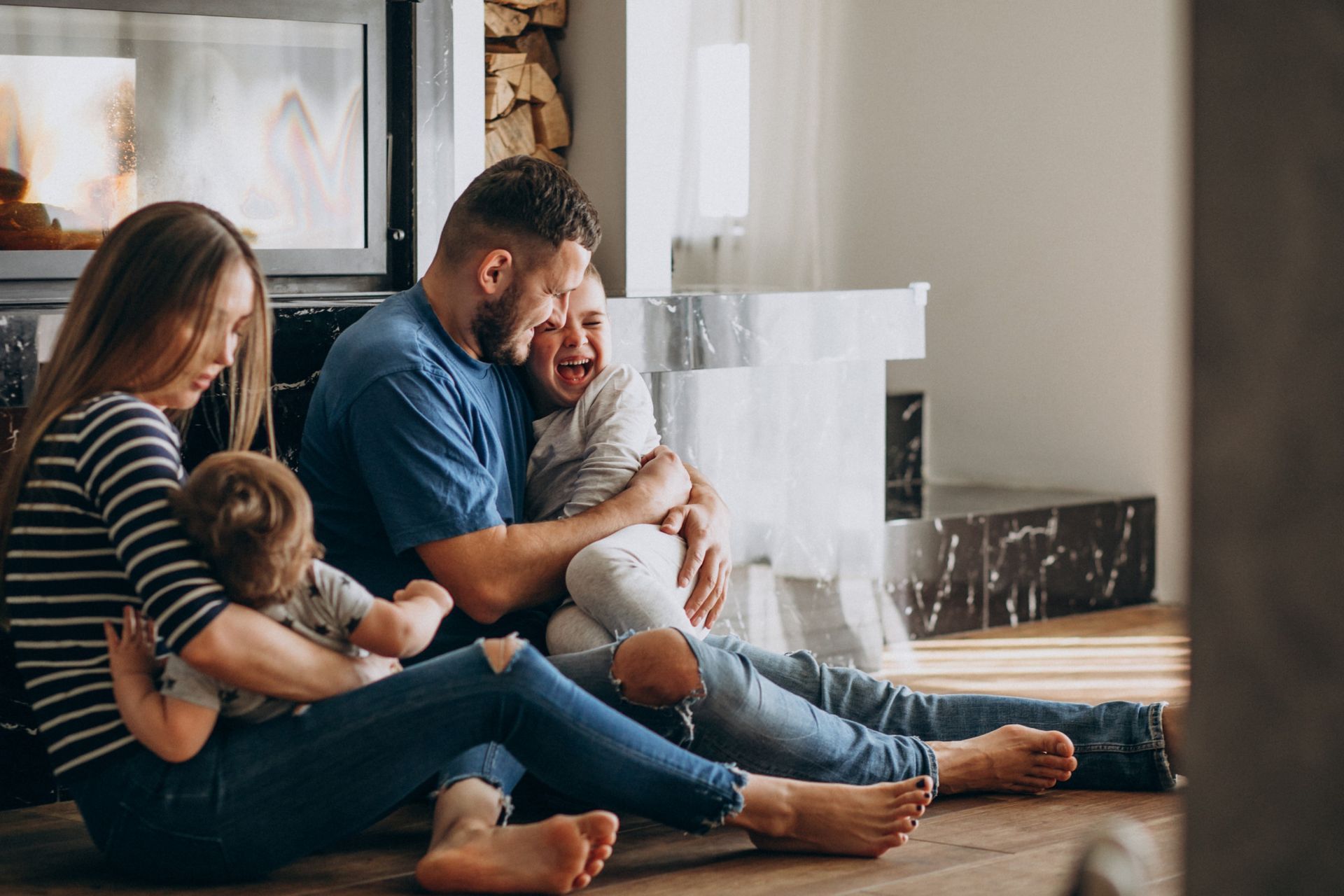 Family of four sits on floor by fireplace, hugging and smiling.