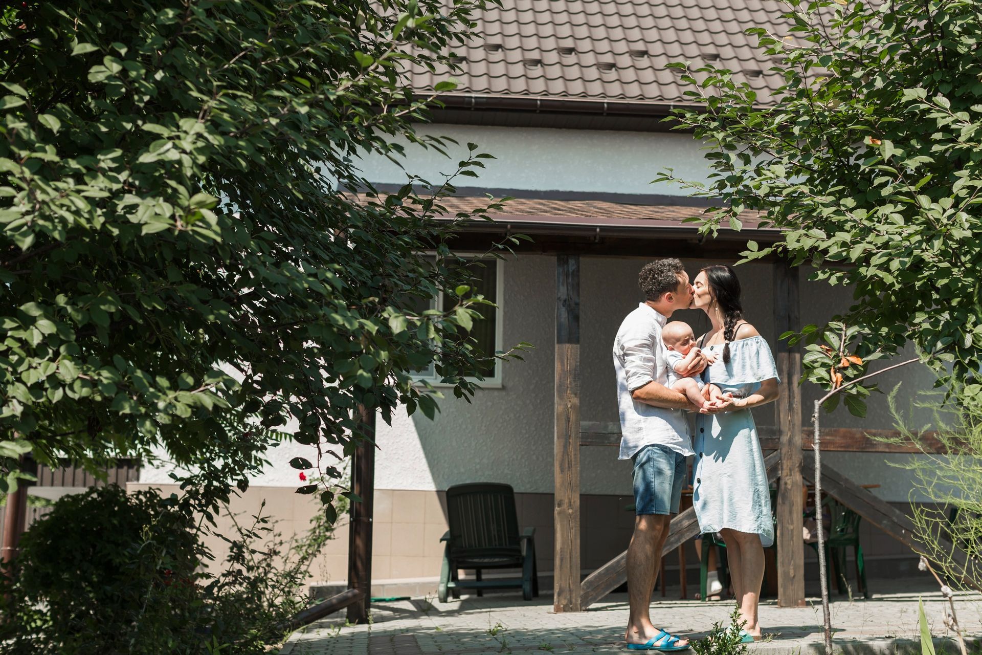 Family kisses outside a house with a porch; man holds baby, woman embraces.