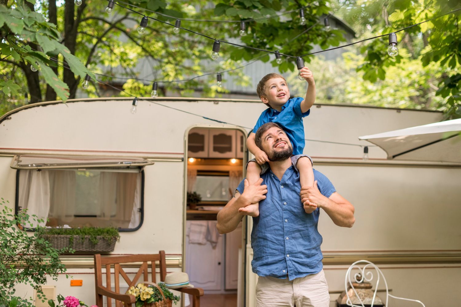 A man carries a child on his shoulders near a camper. The child points up, smiling. Outdoors.
