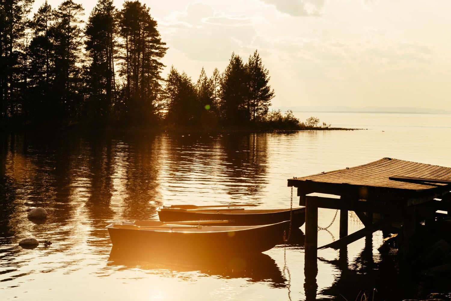Boats docked at a wooden pier on a calm lake, with trees in the background at sunset.