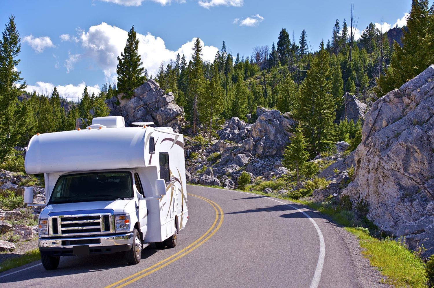 White RV driving on a winding road through a mountainous, wooded landscape under a blue sky.