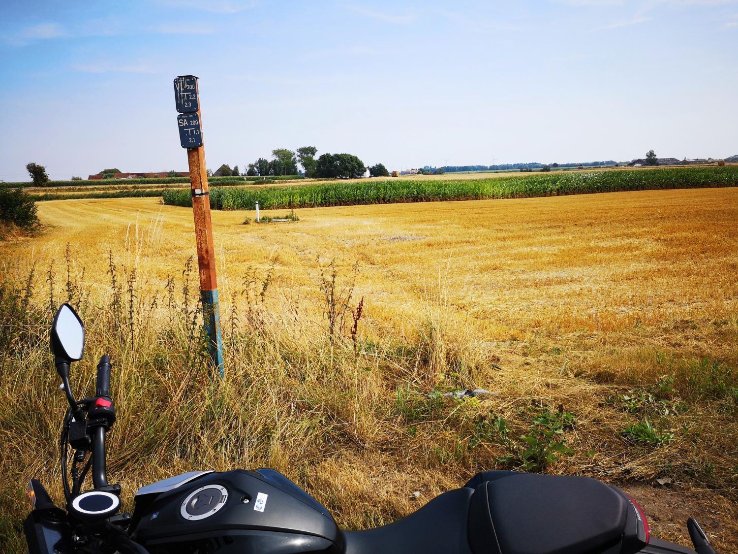 Motorcycle parked by a field of golden wheat, with a wooden pole marker under a blue sky.