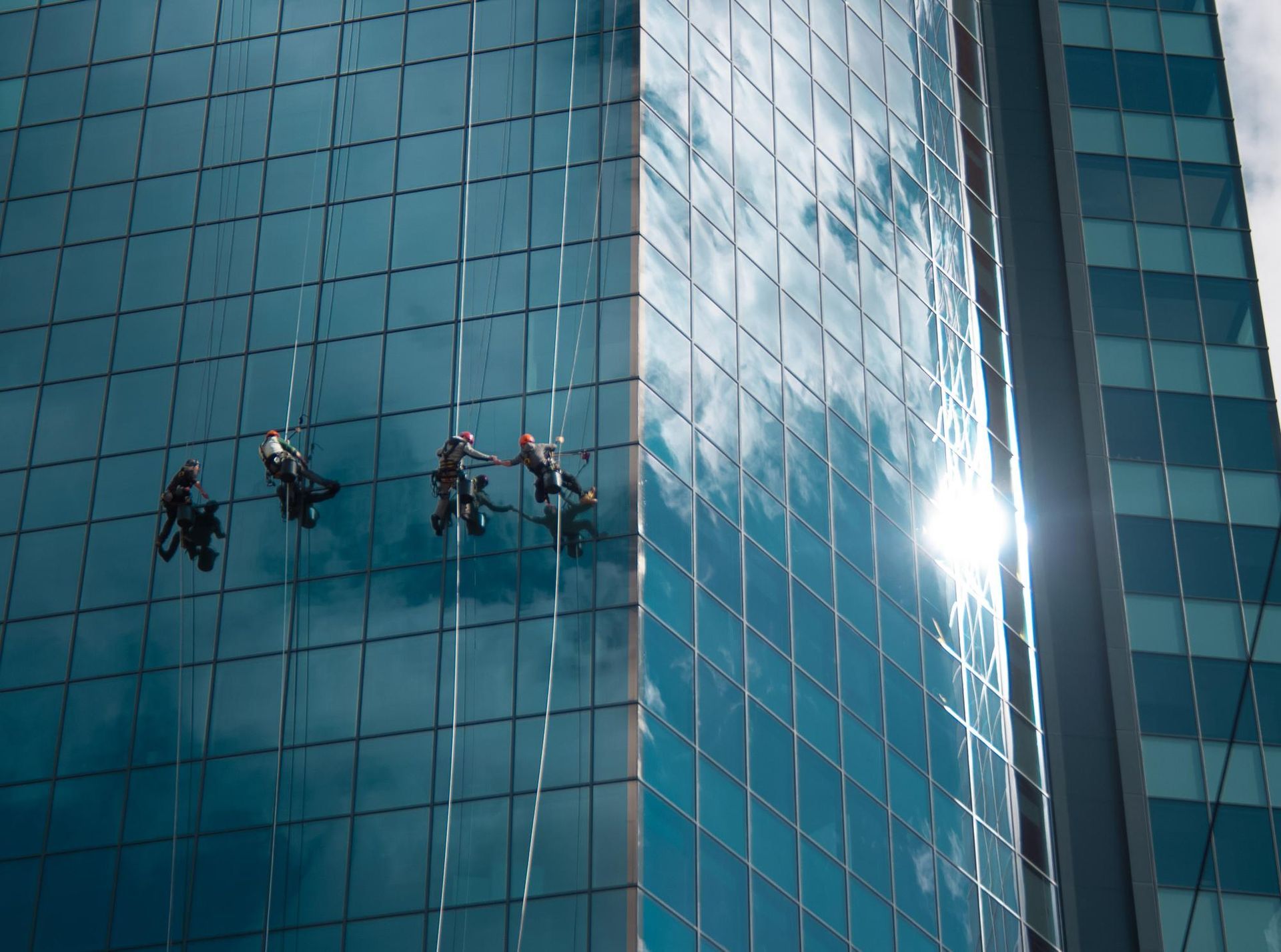 Window washers on a high-rise building, cleaning reflective blue glass under a sunny sky.