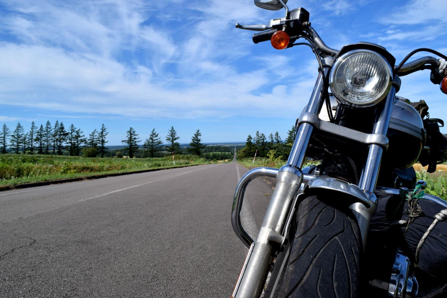 Motorcycle parked on a road, front wheel in focus, with a scenic backdrop of trees and blue sky.