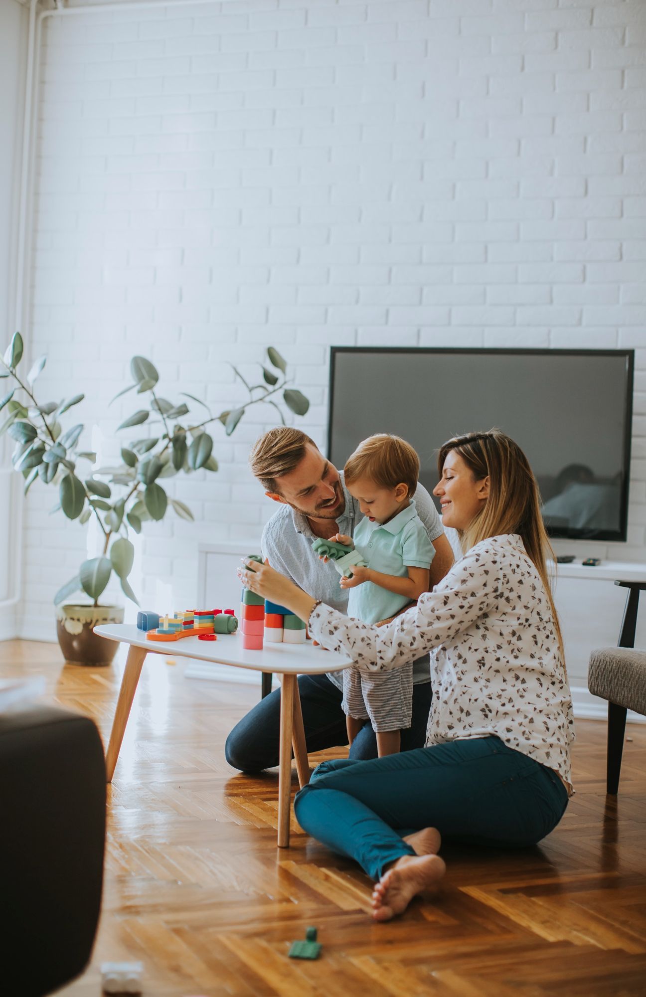 Family playing with a child at a table in a living room; smiling, wood floors, white brick wall.