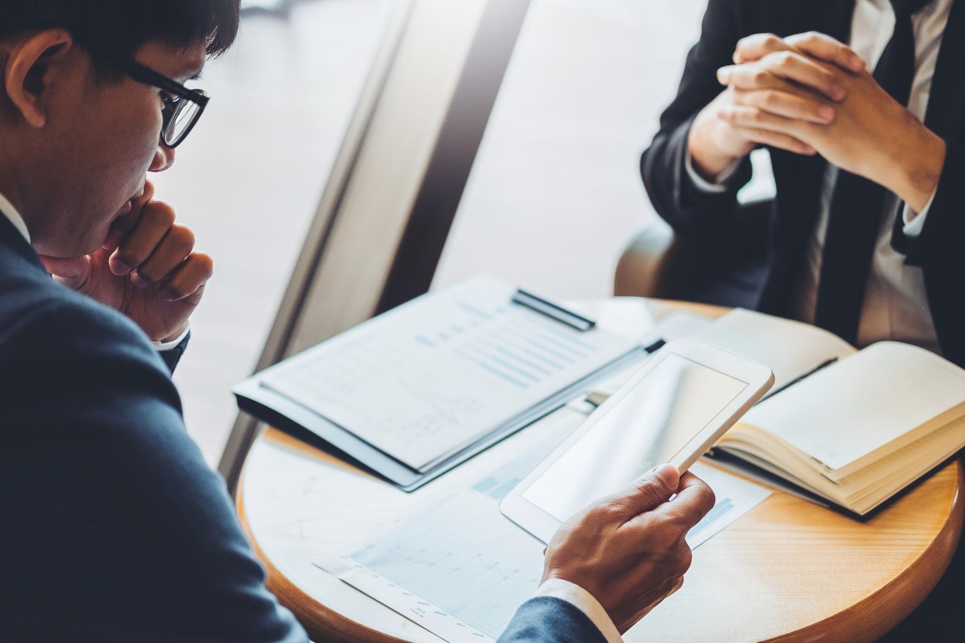 Two people in suits at a round table, reviewing documents, one person has eyeglasses.