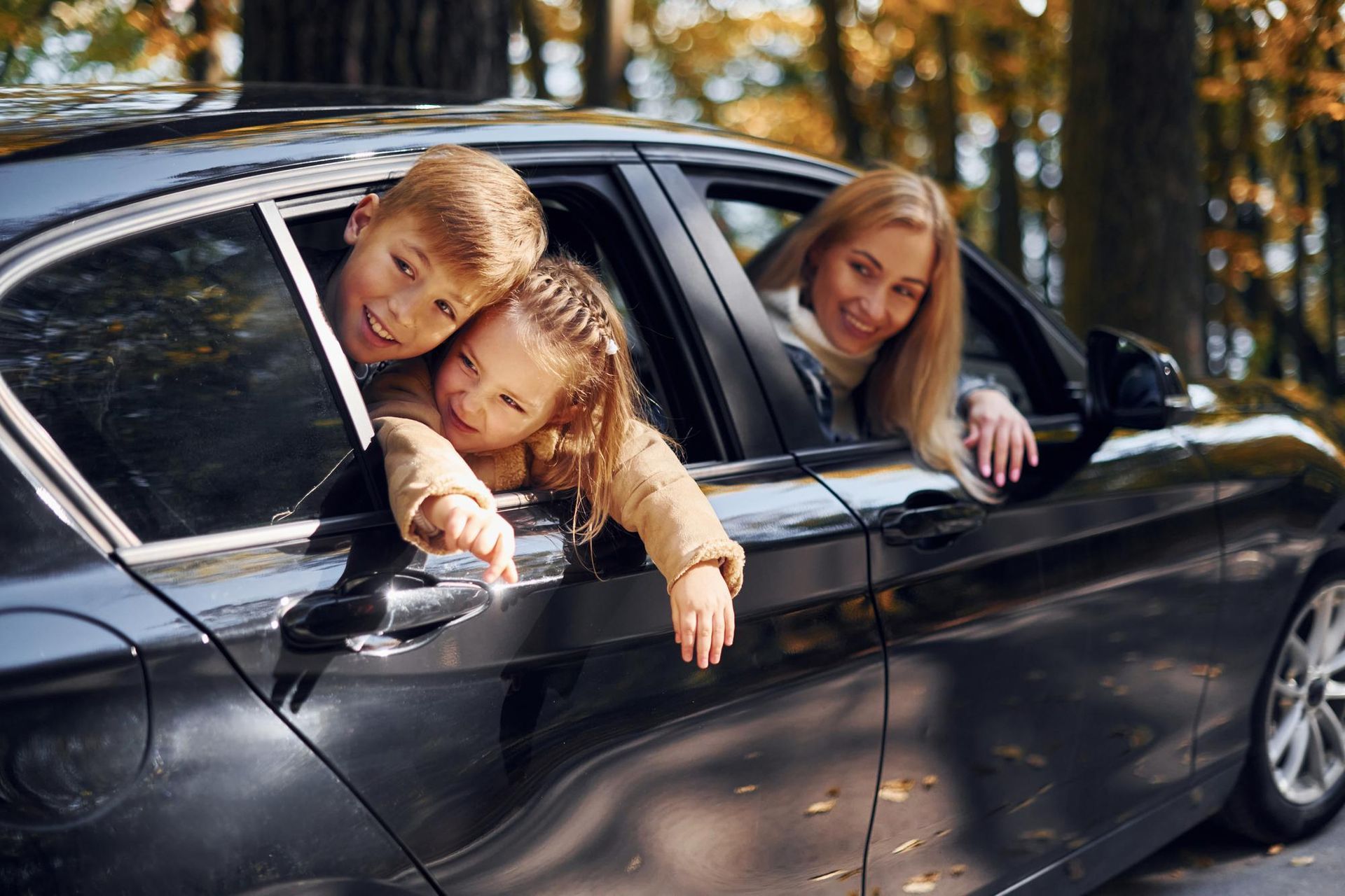 Family in black car, two children smiling and leaning out window, mother smiling from driver's seat, outdoors in autumn.