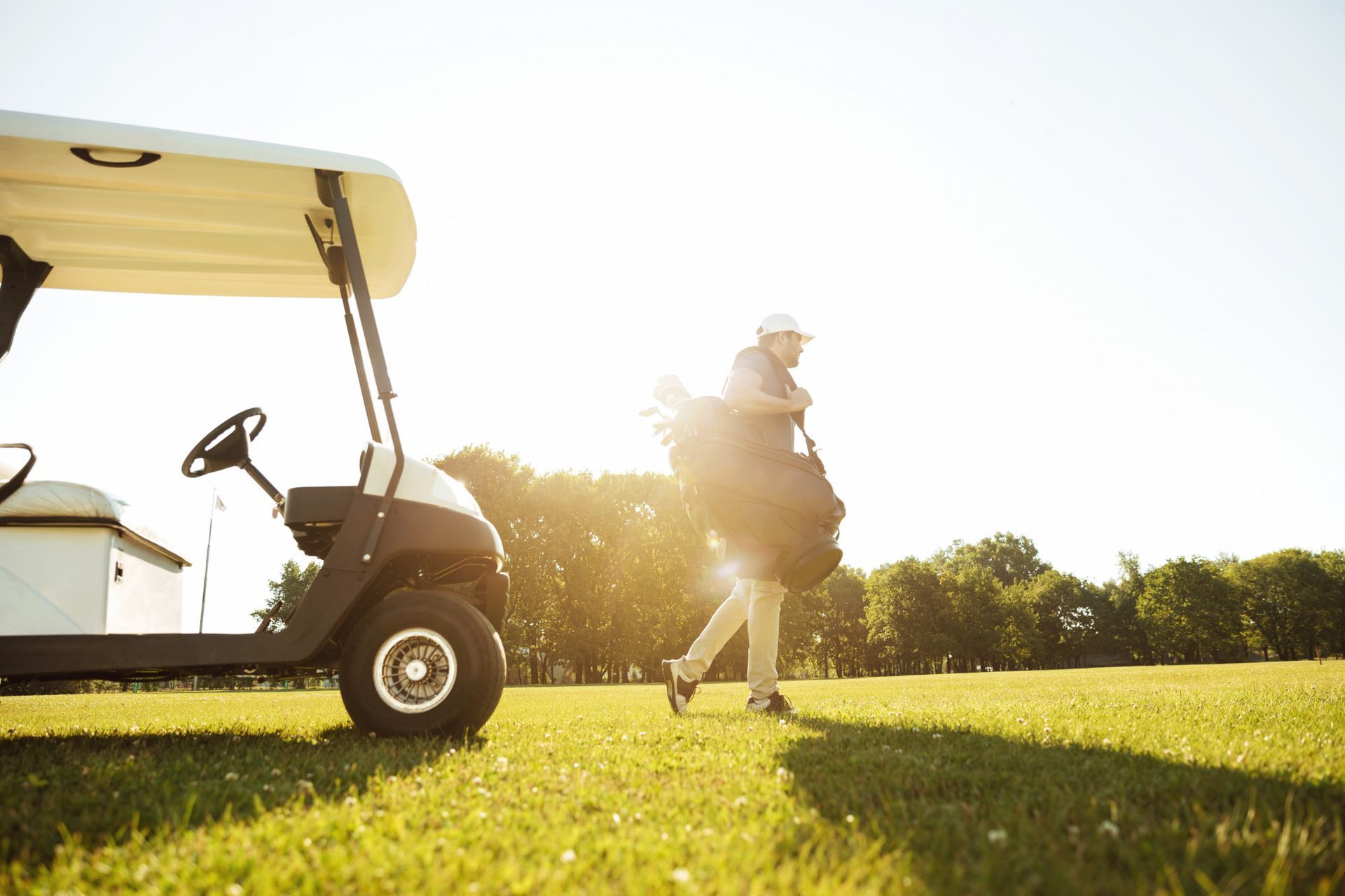 Person carrying golf bag, walking near golf cart on a sunny green golf course.