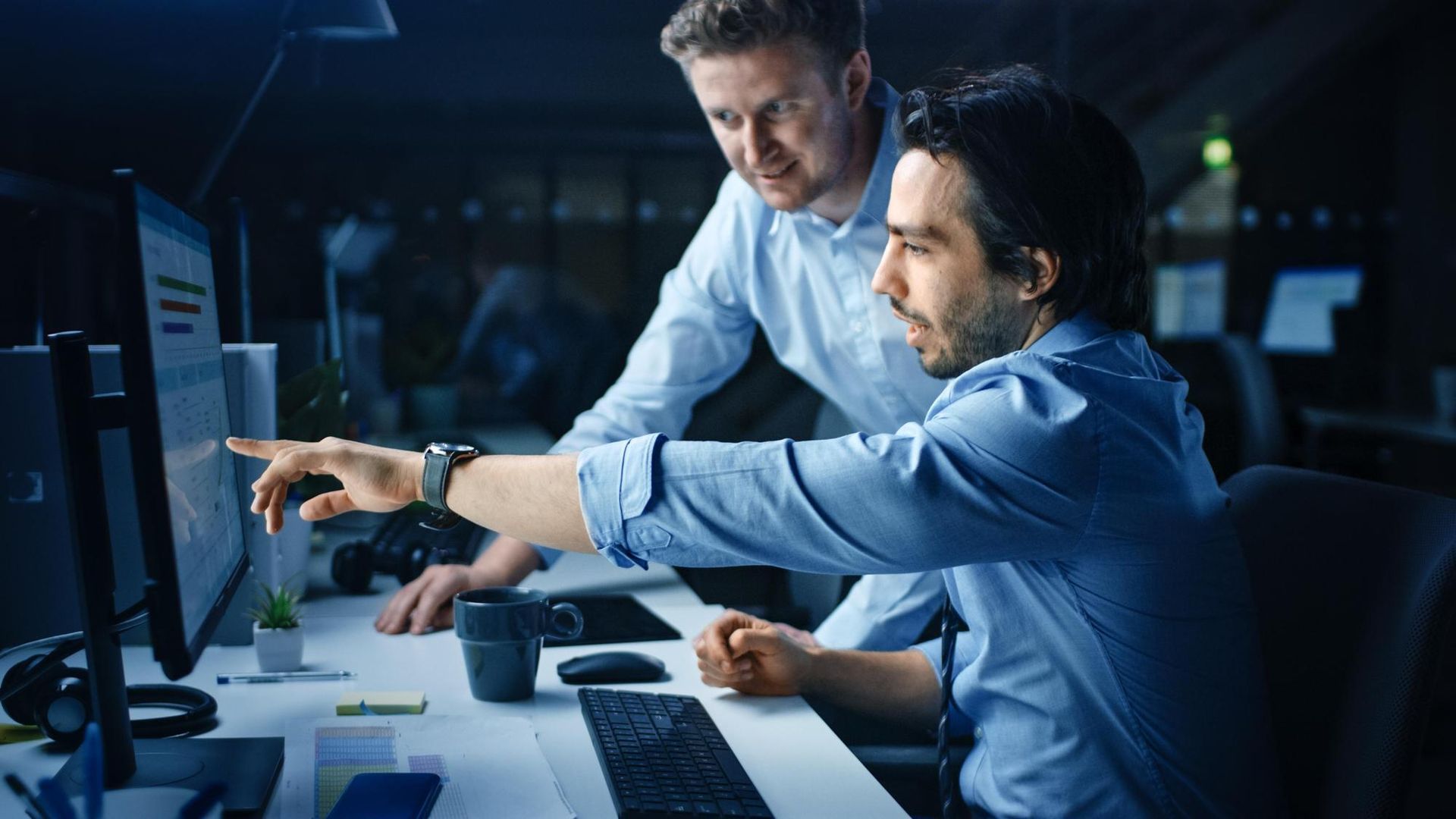 Two men in a dimly lit office look at a computer screen, one pointing.