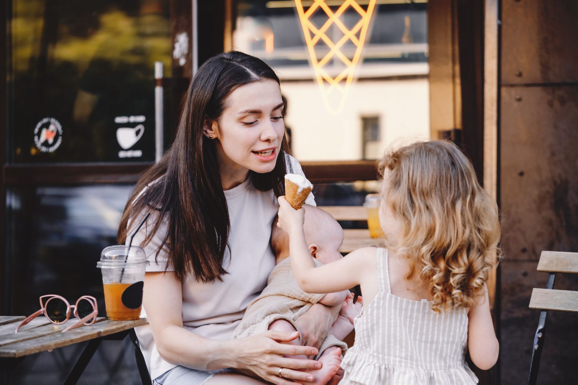 Woman holding baby, sitting outside, and sharing ice cream with toddler.