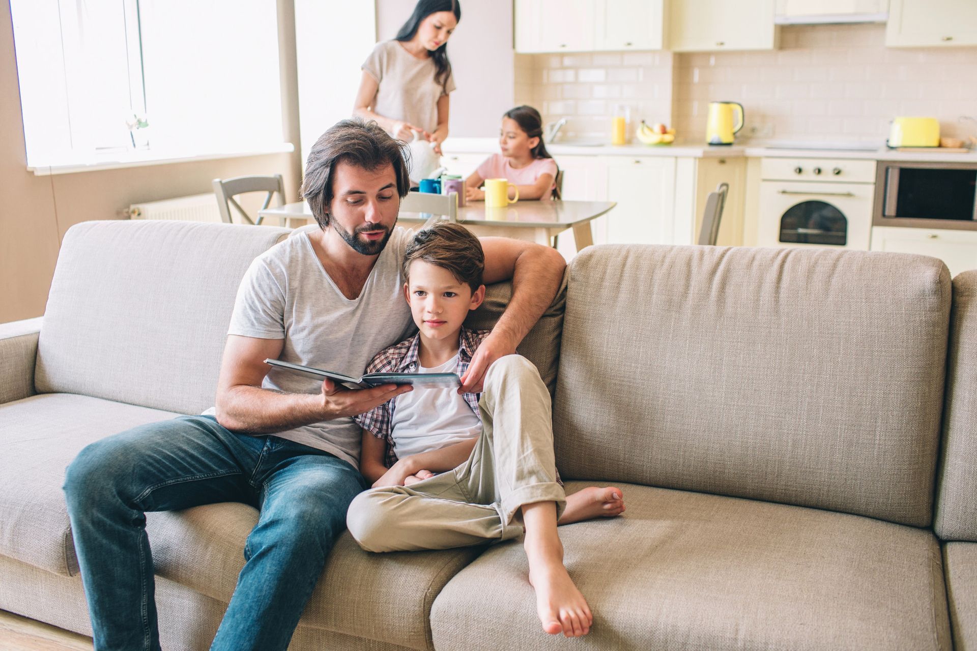 Father and child look at a tablet together on a couch; kitchen and other family members visible in background.