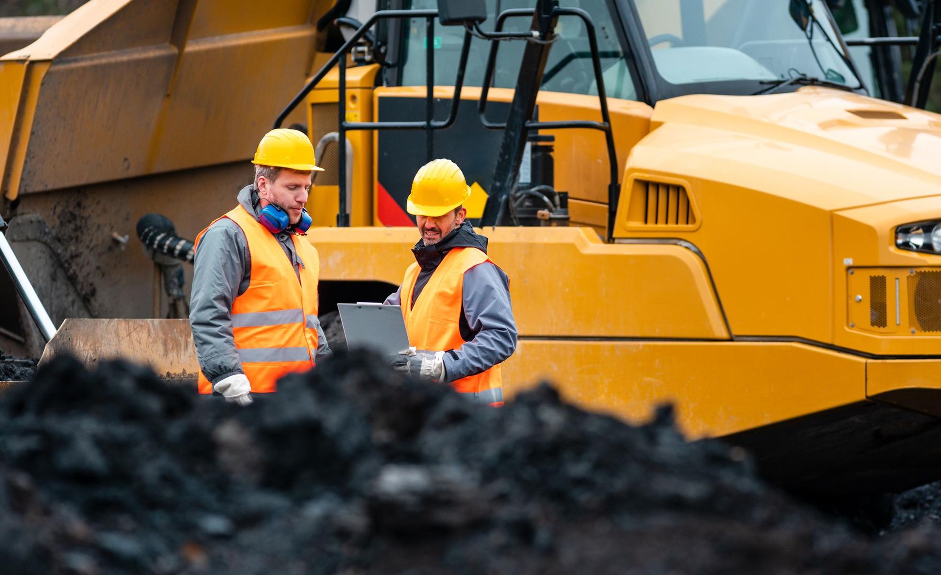 Two workers in safety vests and hard hats reviewing a tablet near a yellow construction vehicle.