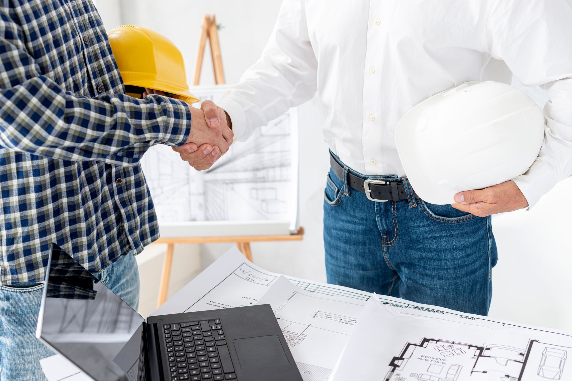 Two construction workers shaking hands at a desk with blueprints and a laptop.